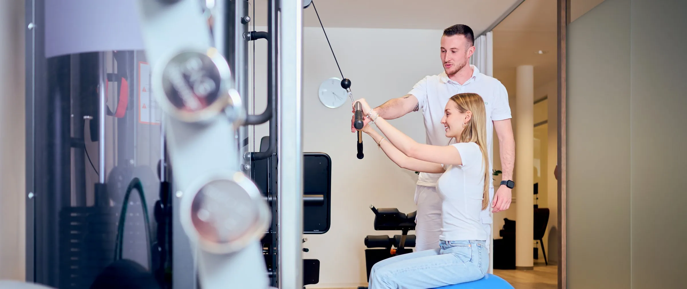Ein Physiotherapeut von SaanoMed begleitet im Trainingsraum eine Patientin, die auf dem Gymnastikball sitzt und am Seilzug zieht